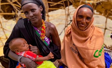 Two women smile at the camera as one of them breastfeeds her baby
