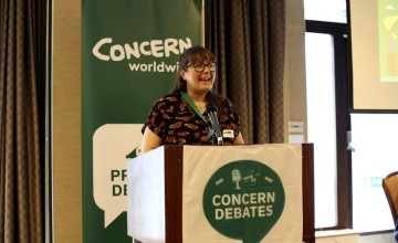 A person stands behind a podium with a microphone, delivering a speech. The podium displays a banner reading 'CONCERN DEBATES' with a microphone and speech bubble icon. Behind them is a 'CONCERN worldwide' banner and part of a projection screen.