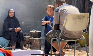 Three people in an outdoor setting: one sitting on the ground to the left holding a spoon, a child in the center interacting with a pot on a makeshift stove, and another person seated on a chair to the right facing away. Various items, including a plastic container, are scattered around them.