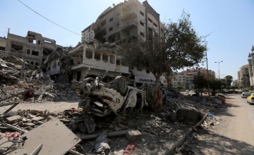 Scene of destruction with collapsed buildings and scattered debris. An overturned car is partially buried under rubble. Damaged structures and a lone tree are visible in the background under a clear daytime sky.