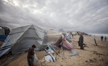 Beach scene with several damaged tents and scattered debris. People are walking and standing among the wreckage, assessing damage or collecting belongings. The sky is overcast, creating a gloomy atmosphere.