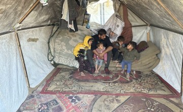 Interior of a makeshift tent with a father and four children sitting on a mattress over patterned rugs. The tent is made of fabric supported by wooden poles, with clothing and blankets hanging inside.