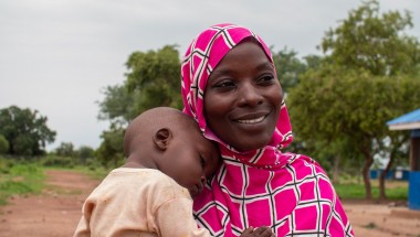 A person wearing a pink and white patterned headscarf holds a child outdoors. In the background, there are trees and a building with a blue roof.