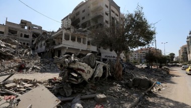 Scene of destruction with collapsed buildings and scattered debris. An overturned car is partially buried under rubble. Damaged structures and a lone tree are visible in the background under a clear daytime sky.