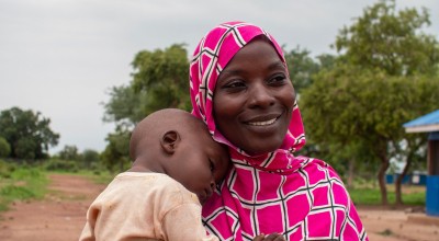 A person wearing a pink and white patterned headscarf holds a child outdoors. In the background, there are trees and a building with a blue roof.